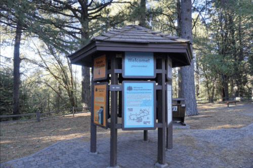 A wooden information kiosk in a forested area, featuring colorful welcome signs and maps.