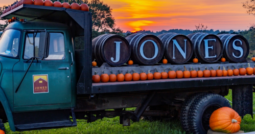 A vintage truck with pumpkins and barrels, set against a colorful sunset. The truck displays the name "JONES."