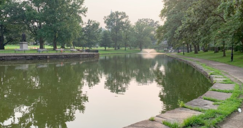 A serene park scene featuring a calm pond with a fountain, surrounded by trees and a walking path.