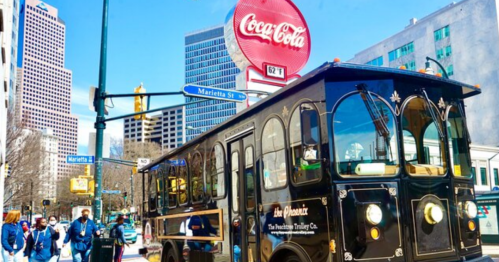 A vintage trolley bus stops near a Coca-Cola sign in a bustling city street with pedestrians walking by.