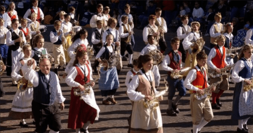 A lively parade scene with children and adults in traditional costumes, playing instruments and marching together.