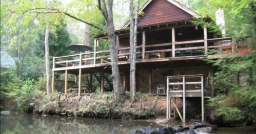 A wooden cabin with a deck, surrounded by trees, overlooking a calm river.