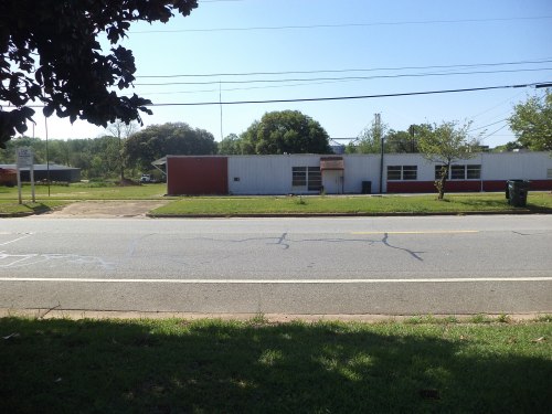 A view of a quiet street with a building in the background, surrounded by trees and power lines under a clear blue sky.