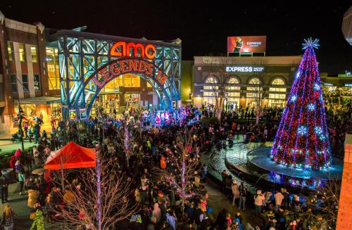 A bustling outdoor shopping area at night, featuring a large Christmas tree and crowds of people celebrating the holiday season.