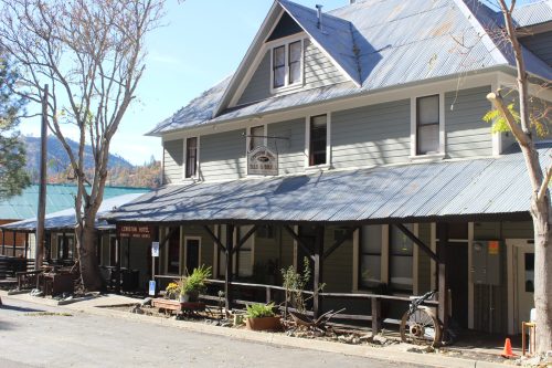 A large, two-story gray building with a metal roof, featuring a porch and signs, surrounded by trees and mountains.