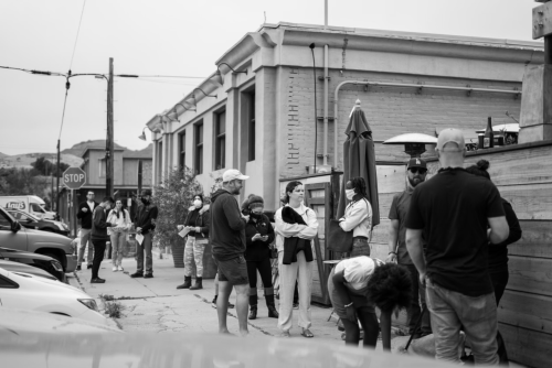 A black and white image of a crowd waiting outside a building, some wearing masks, with parked cars in the foreground.