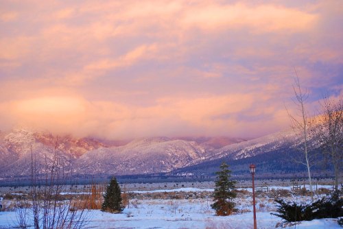 Snow-covered mountains under a colorful sunset sky, with trees and a field in the foreground.