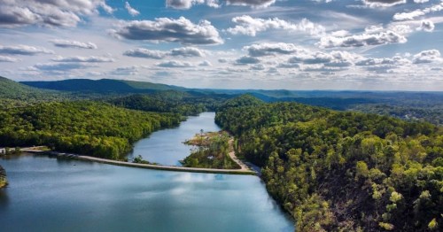 Aerial view of a serene lake surrounded by lush green hills and scattered clouds in a blue sky.