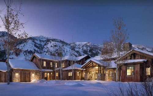 A cozy mountain lodge surrounded by snow-covered trees and mountains, illuminated at dusk.
