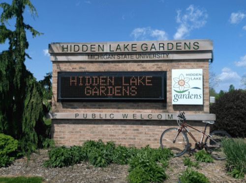 Sign for Hidden Lake Gardens at Michigan State University, featuring a bike and welcoming visitors.