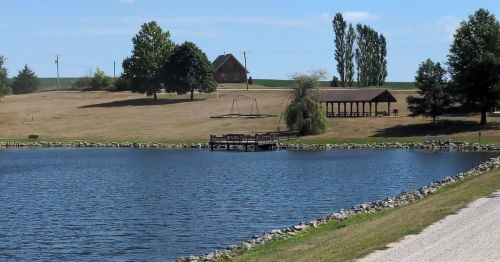 A serene landscape featuring a calm lake, a wooden dock, a house, and trees under a clear blue sky.