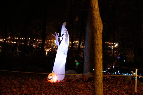 A ghostly figure stands in a dark forest, with a carved pumpkin and colorful lights in the background.