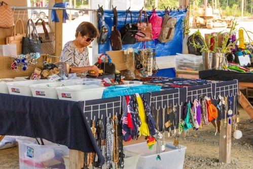A vendor arranges jewelry and accessories at a market stall, surrounded by bags and colorful items for sale.