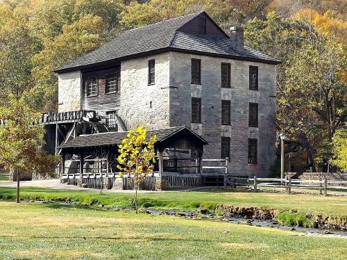 Historic stone mill building with a waterwheel, surrounded by trees and a grassy area.