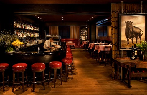 A dimly lit restaurant bar with red stools, a bartender, and a large painting of a cow on the wall.