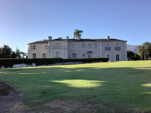 A large, elegant mansion with a manicured lawn and palm trees, set against a clear blue sky.