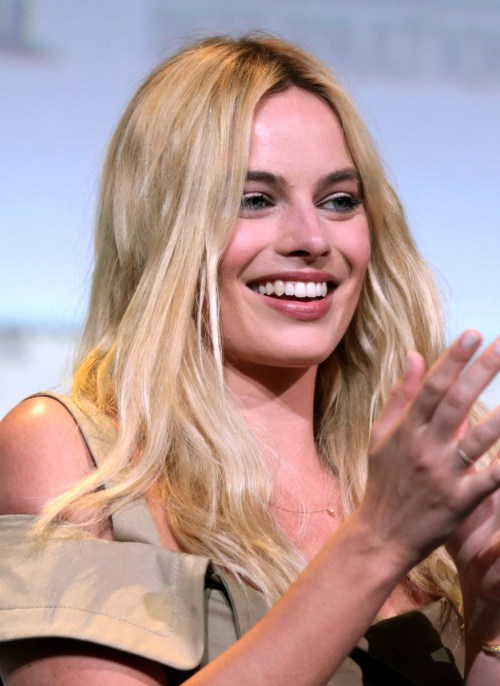 A smiling woman with long blonde hair, wearing a light-colored dress, gestures while speaking at an event.
