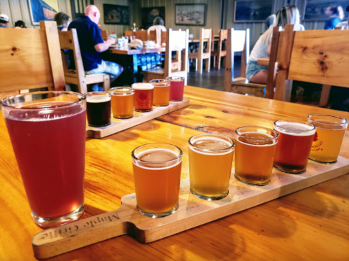 A wooden table with two flight trays of various craft beers and a glass of pink beer in a rustic restaurant setting.