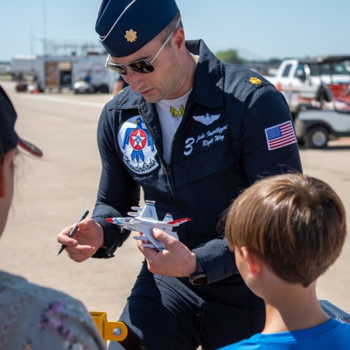 A pilot in a blue flight suit signs an airplane model for two children at an airshow.