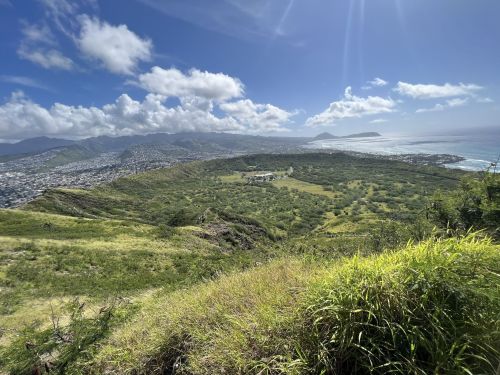 A panoramic view of a lush green landscape with mountains and coastline under a bright blue sky with scattered clouds.