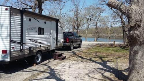 A parked RV and truck by a serene lake, surrounded by bare trees and a gravel path.