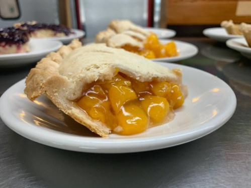 A slice of peach pie on a white plate, featuring a golden filling and a flaky crust.