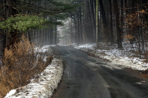 A misty road winding through a forest, with patches of snow and bare trees on either side.