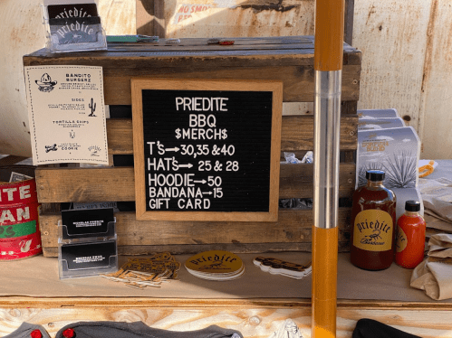 A wooden display with a sign listing BBQ merchandise prices, including T-shirts, hats, hoodies, and gift cards.