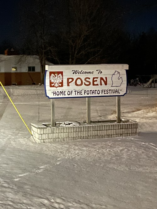 Welcome sign for Posen, Michigan, stating "Home of the Potato Festival," surrounded by snow at night.