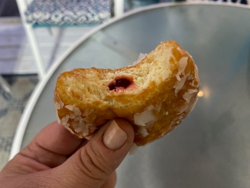 A hand holding a partially eaten glazed donut with a visible filling of red fruit jam.