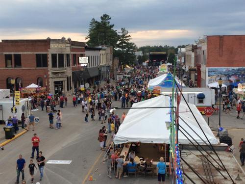 A bustling street fair with crowds, food tents, and vendors under a cloudy sky, lined by historic buildings.