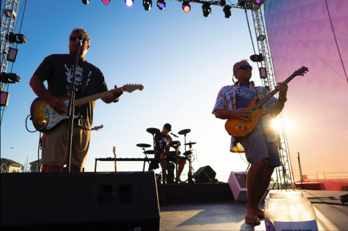 A band performs on stage at sunset, with a guitarist and vocalist in the foreground and a drummer in the background.