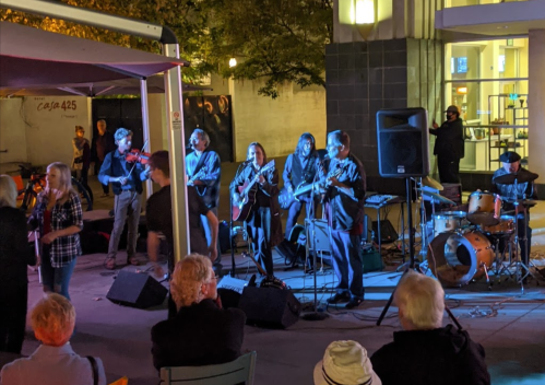 A live band performs outdoors at night, with musicians playing guitars and drums, while an audience enjoys the show.