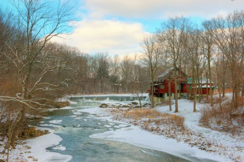 A snowy landscape with a river, a waterfall, and a red cabin surrounded by bare trees under a blue sky.
