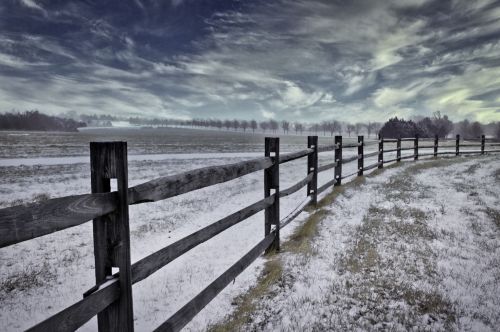 A snow-covered field with a wooden fence curving through the landscape under a cloudy sky.