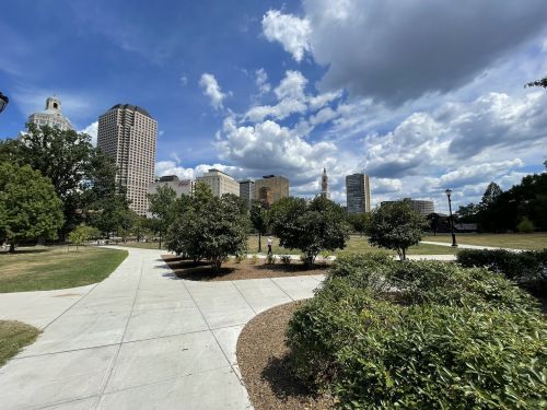 A park pathway lined with greenery, leading to a city skyline under a partly cloudy sky.