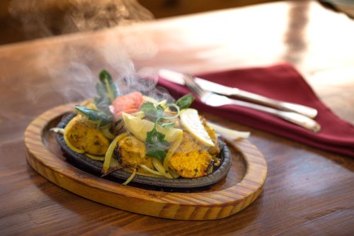 Sizzling platter of grilled vegetables and spices, garnished with fresh herbs and lemon, on a wooden serving board.