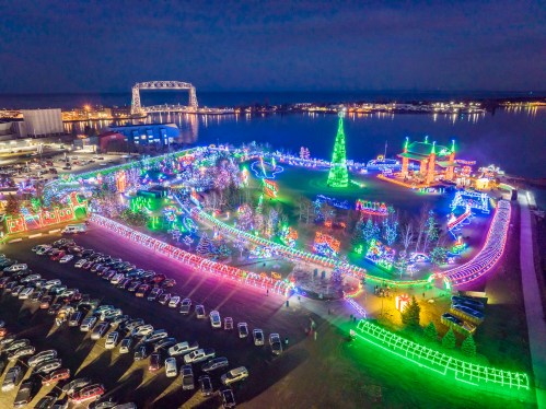 Aerial view of a festive park illuminated with colorful lights, featuring a large Christmas tree and a river in the background.