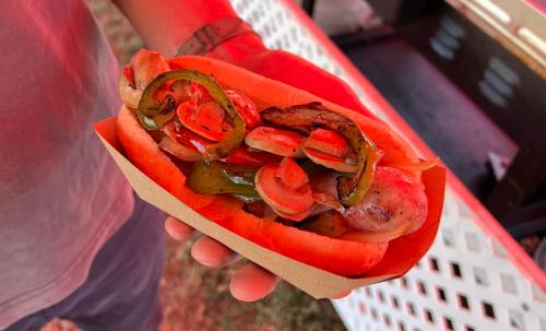 A hand holding a hot dog topped with grilled onions and green peppers in a paper tray.