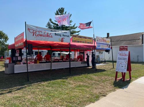 A food stand with red tents and banners, serving Nowicki's products, set up in a grassy area with an American flag.