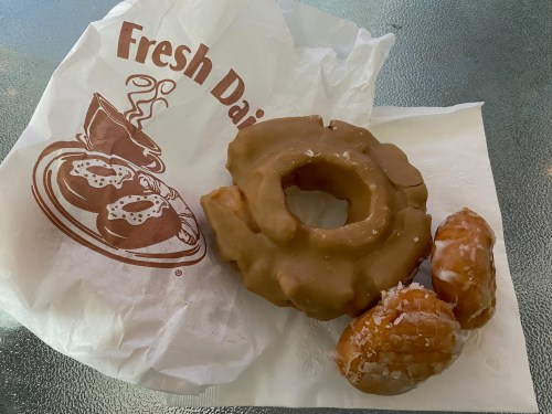 A glazed donut and two small pastries on a paper napkin with a logo, resting on a glass surface.