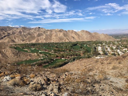 A scenic view of a golf course nestled in a valley surrounded by mountains under a blue sky with scattered clouds.