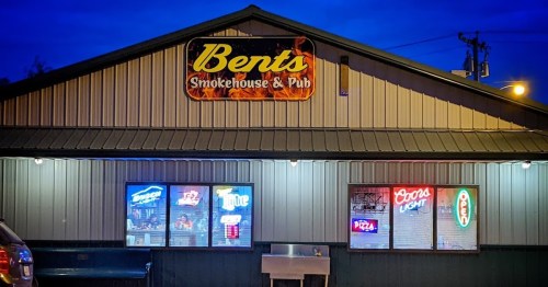 Exterior of Bents Smokehouse & Pub at night, featuring a sign and illuminated windows displaying menu items and drinks.