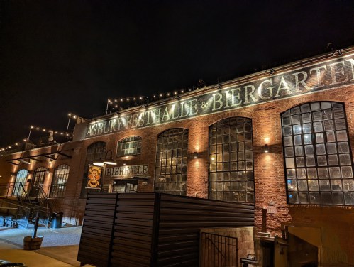 A brick building at night with illuminated signage reading "Asbury Hall & Biergarten" and string lights above.