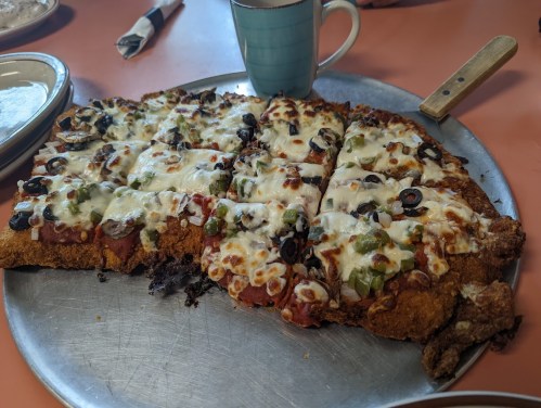 A large, cheesy pizza with black olives and green peppers on a metal tray, alongside a mug on a pink table.