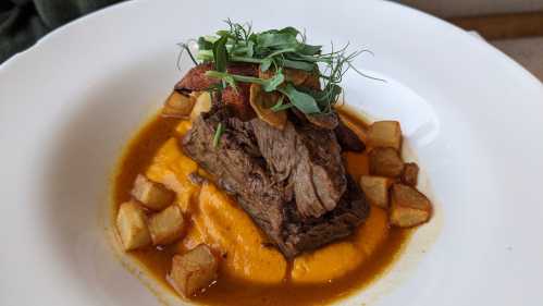 A plate of tender beef served on a bed of orange sauce, garnished with greens and crispy potatoes.