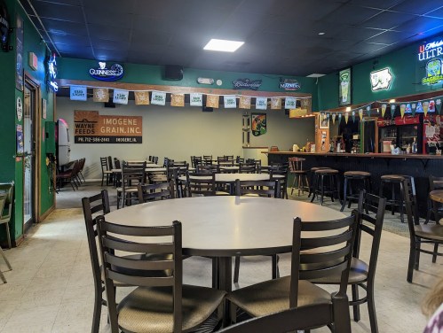 A spacious bar interior with empty tables and chairs, green walls, and a long counter with various drink signs.