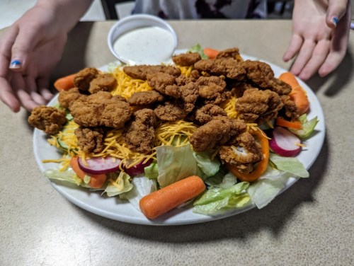 A large plate of salad topped with fried chicken, cheese, and assorted vegetables, with a side of dressing.