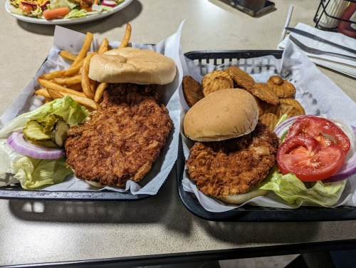 Two fried chicken sandwiches with lettuce, tomato, and pickles, served with fries and potato wedges on a tray.