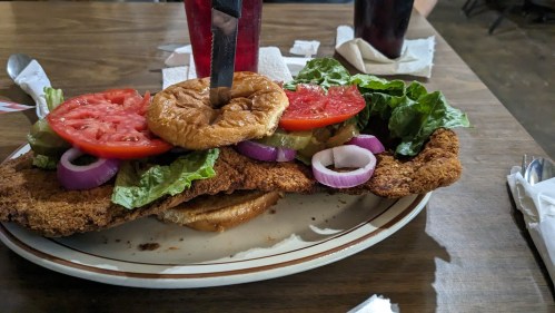 A large fried chicken sandwich topped with lettuce, tomatoes, pickles, and onions, served on a plate.